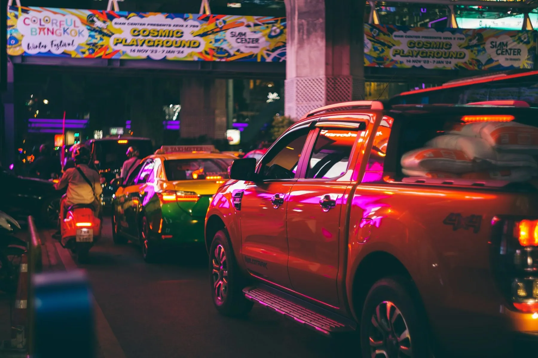 A red pickup, yellow taxis and scooters packed together in neon-lit Bangkok traffic after dark