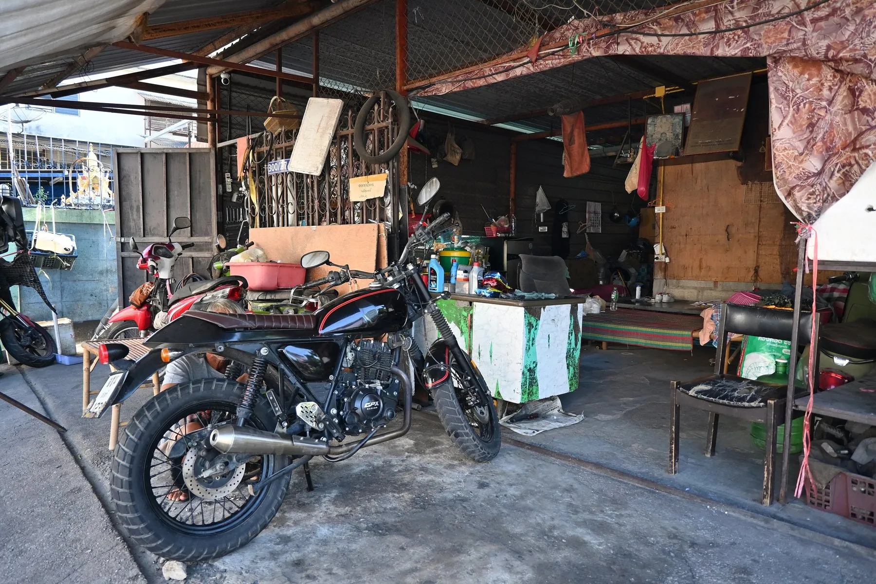 A black dual-sport motorbike on its stand outside a small open Thai workshop hung with tools and spare parts