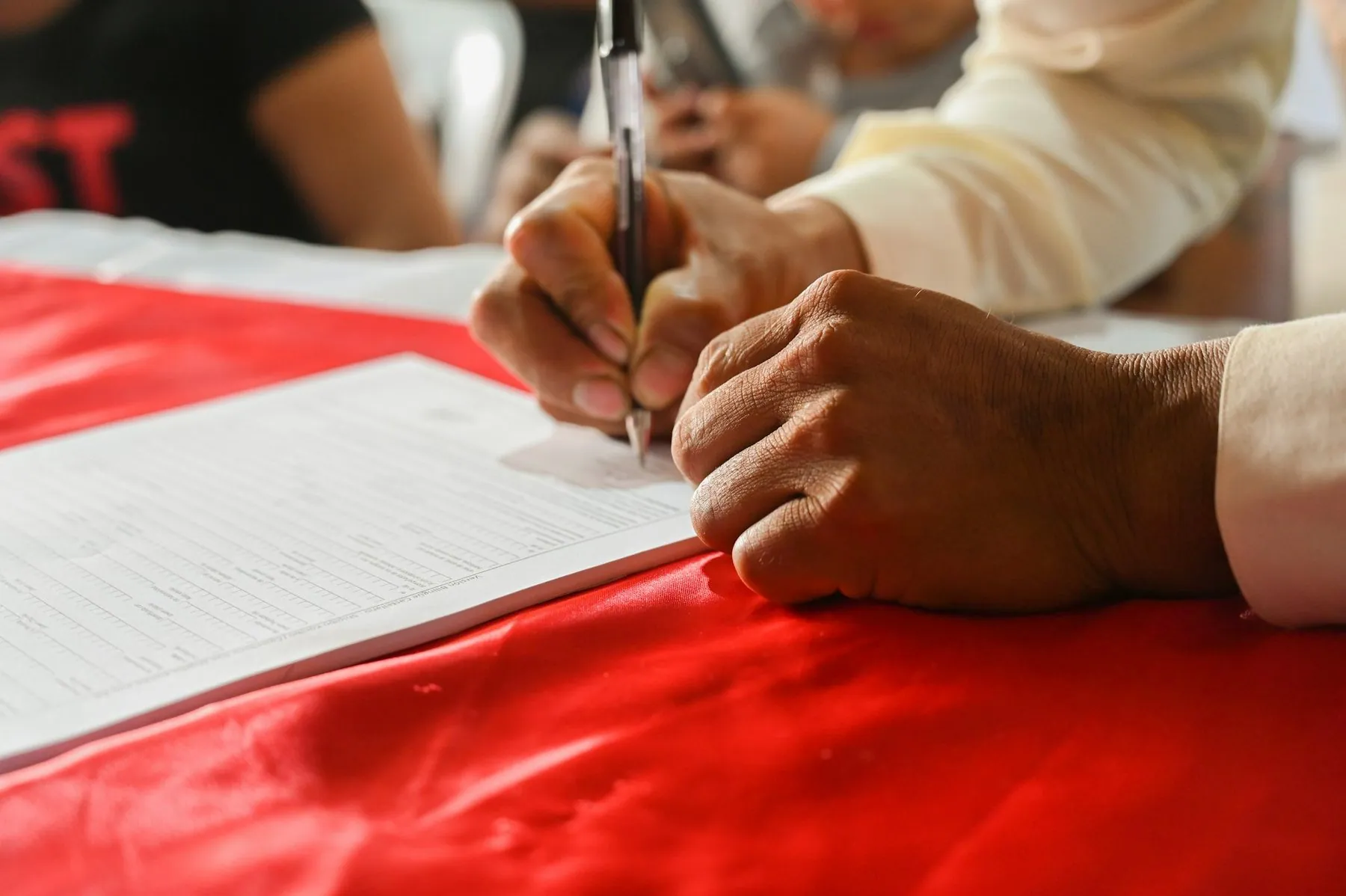 Two pairs of hands filling in printed forms side by side at a table covered with red cloth