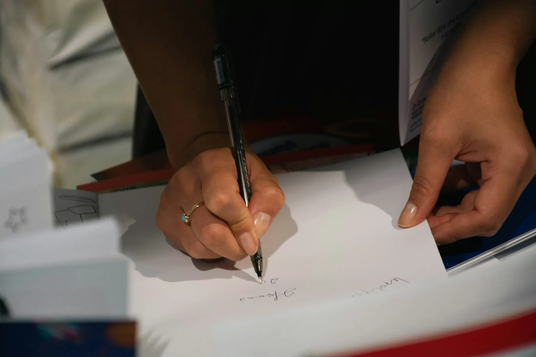 Two hands signing and steadying a printed document on a desk under a hard light