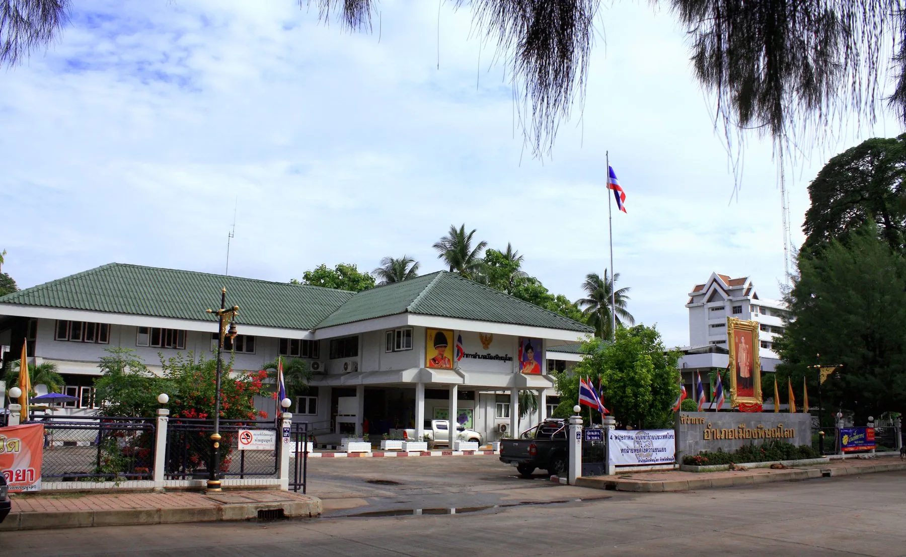 The white district office at Mueang Phitsanulok behind its forecourt, the Thai flag on a pole and vehicles parked along the frontage