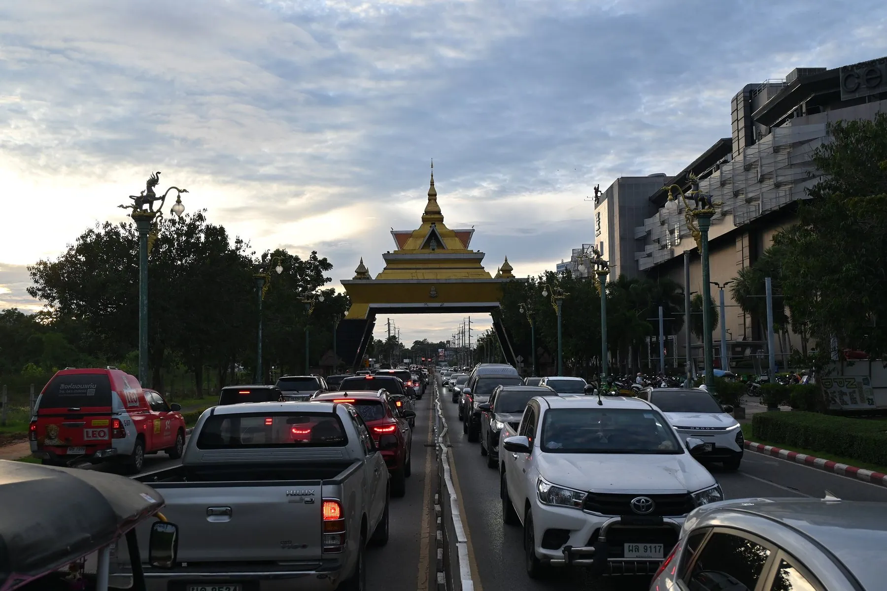 Cars and pickups queueing in several lanes beneath a gilded Thai ceremonial arch as the sun goes down