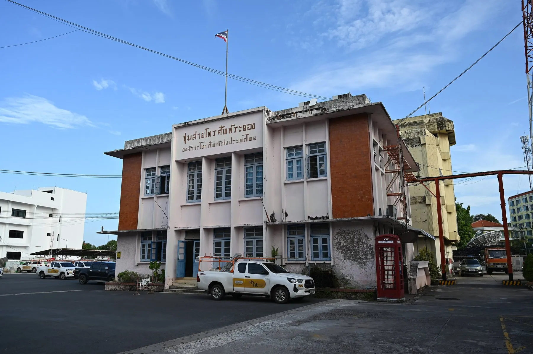 A two-storey Thai government office in orange and white with the national flag on the roof and pickups parked across the forecourt