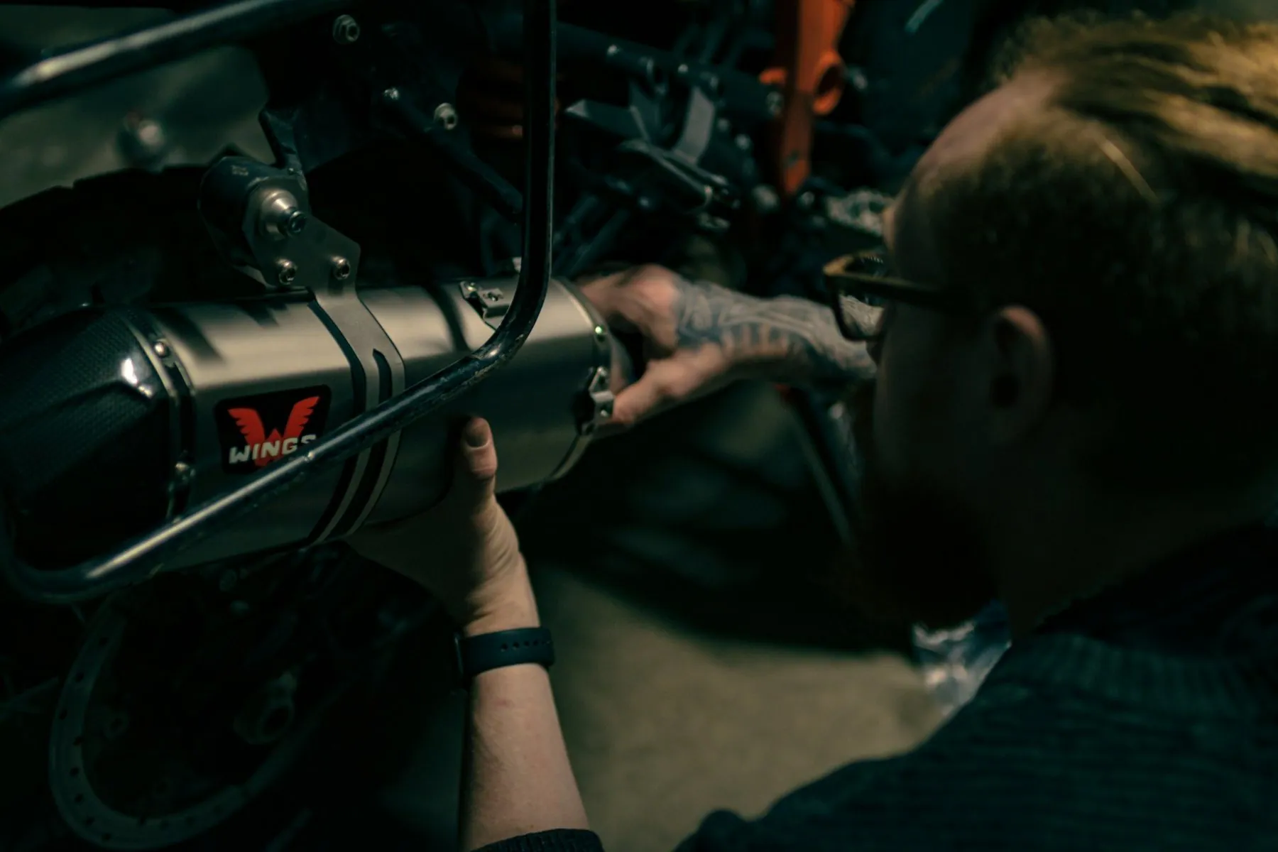 A man leaning over a motorbike in a dim garage, turning a spanner on the engine