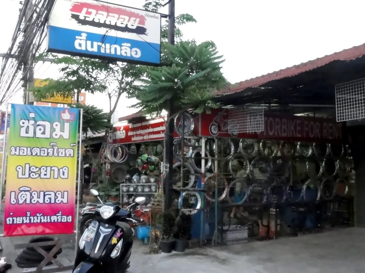 A roadside Thai motorbike shop with a yellow Thai-script banner advertising repairs, tyres and oil changes, wheels racked behind