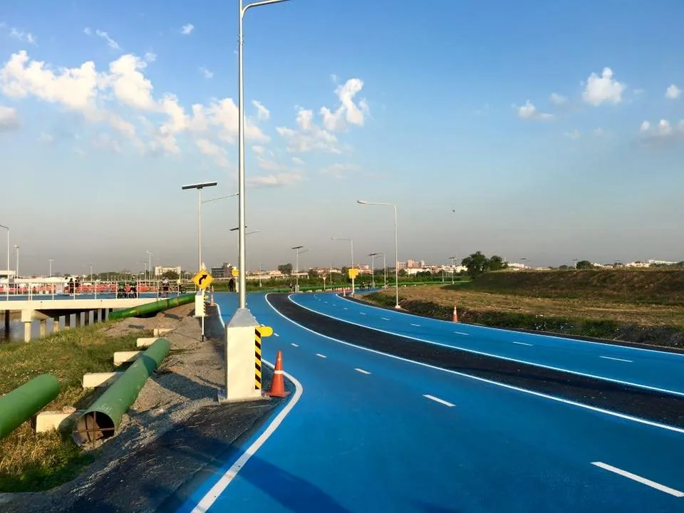 The blue asphalt of the Sky Lane cycling track curving away between crash barriers and lamp posts on the Suvarnabhumi Airport perimeter