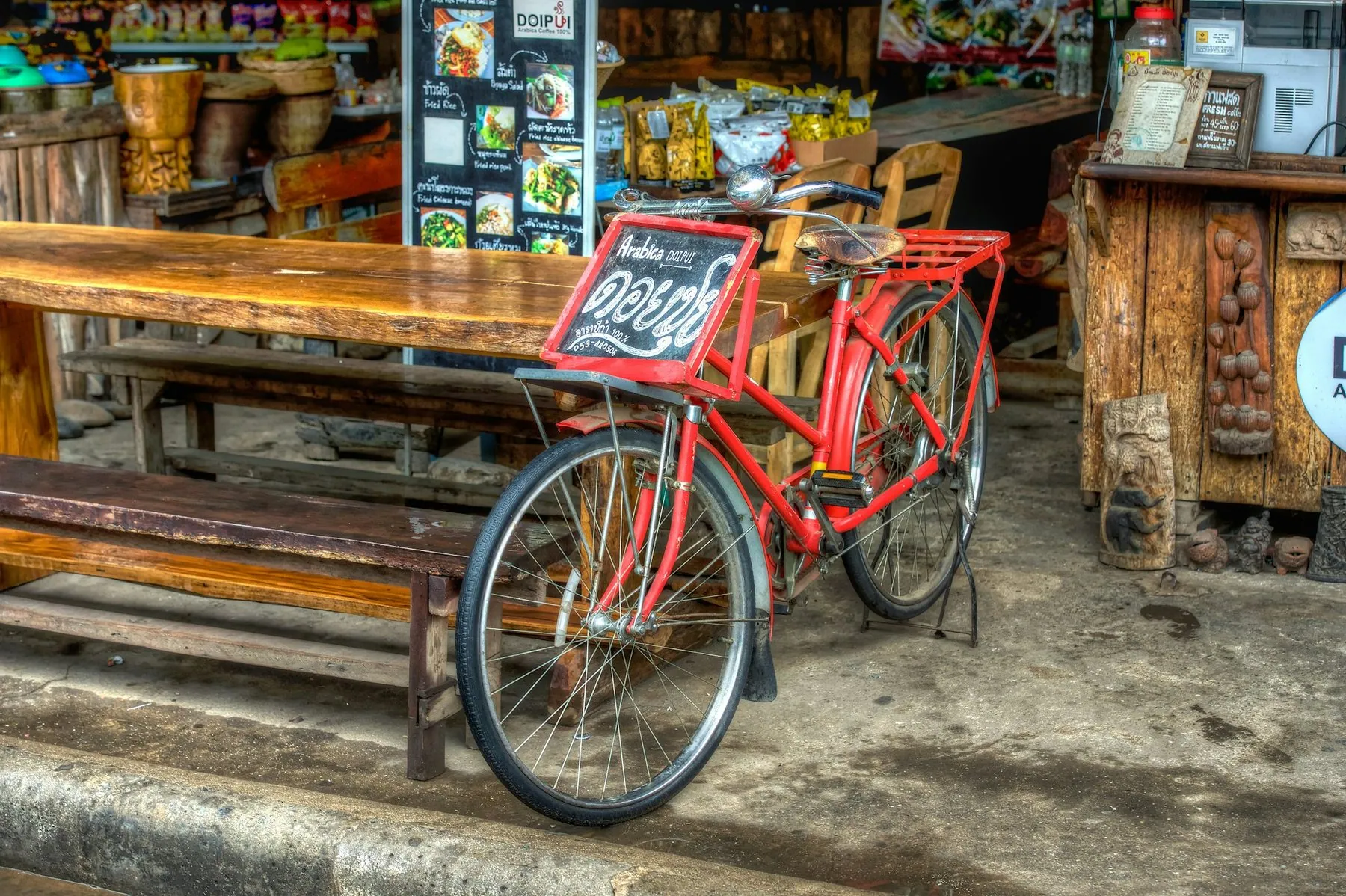 A red delivery bicycle carrying a chalkboard coffee sign in Thai on its front rack, parked beside long wooden benches outside a stall in Chiang Mai