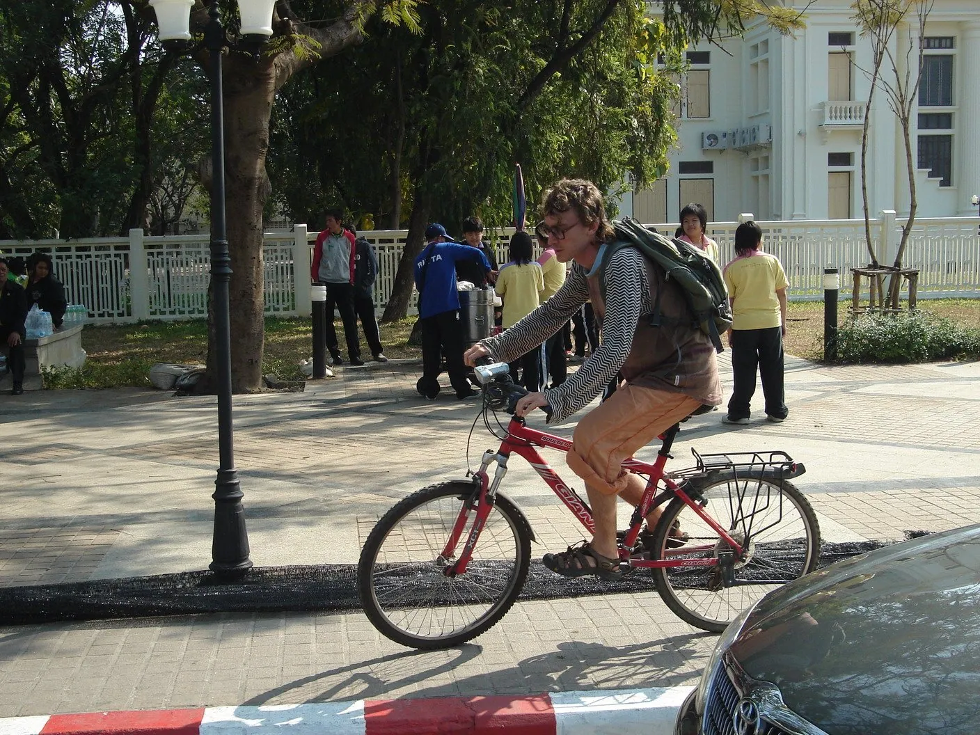 A foreign visitor with a large green rucksack riding a red rack-equipped touring bicycle along a tree-lined street in Chiang Mai