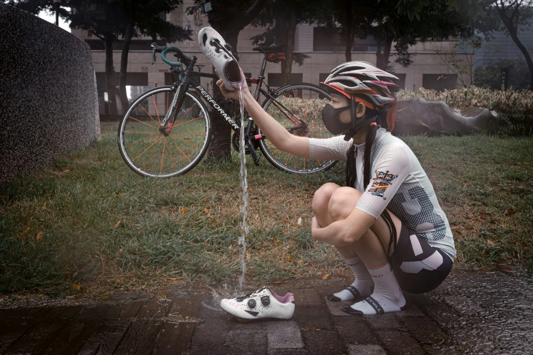 A cyclist in helmet, mitts and stiff-soled shoes crouching beside a bike to refill a bottle from a tap