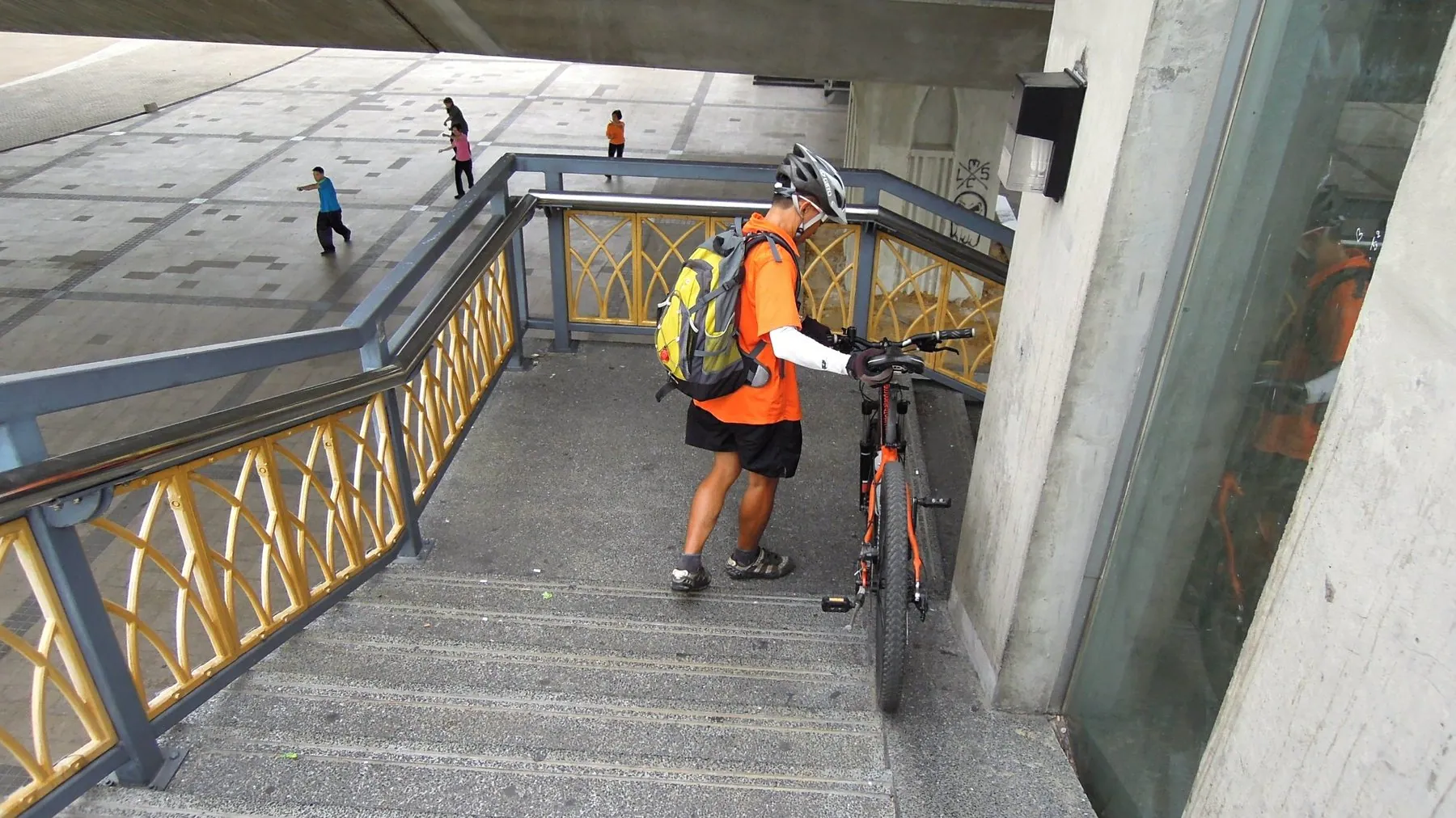 A cyclist in a high-visibility jersey pushing his bike up the gold-railed steps of the Rama VIII Bridge in Bangkok