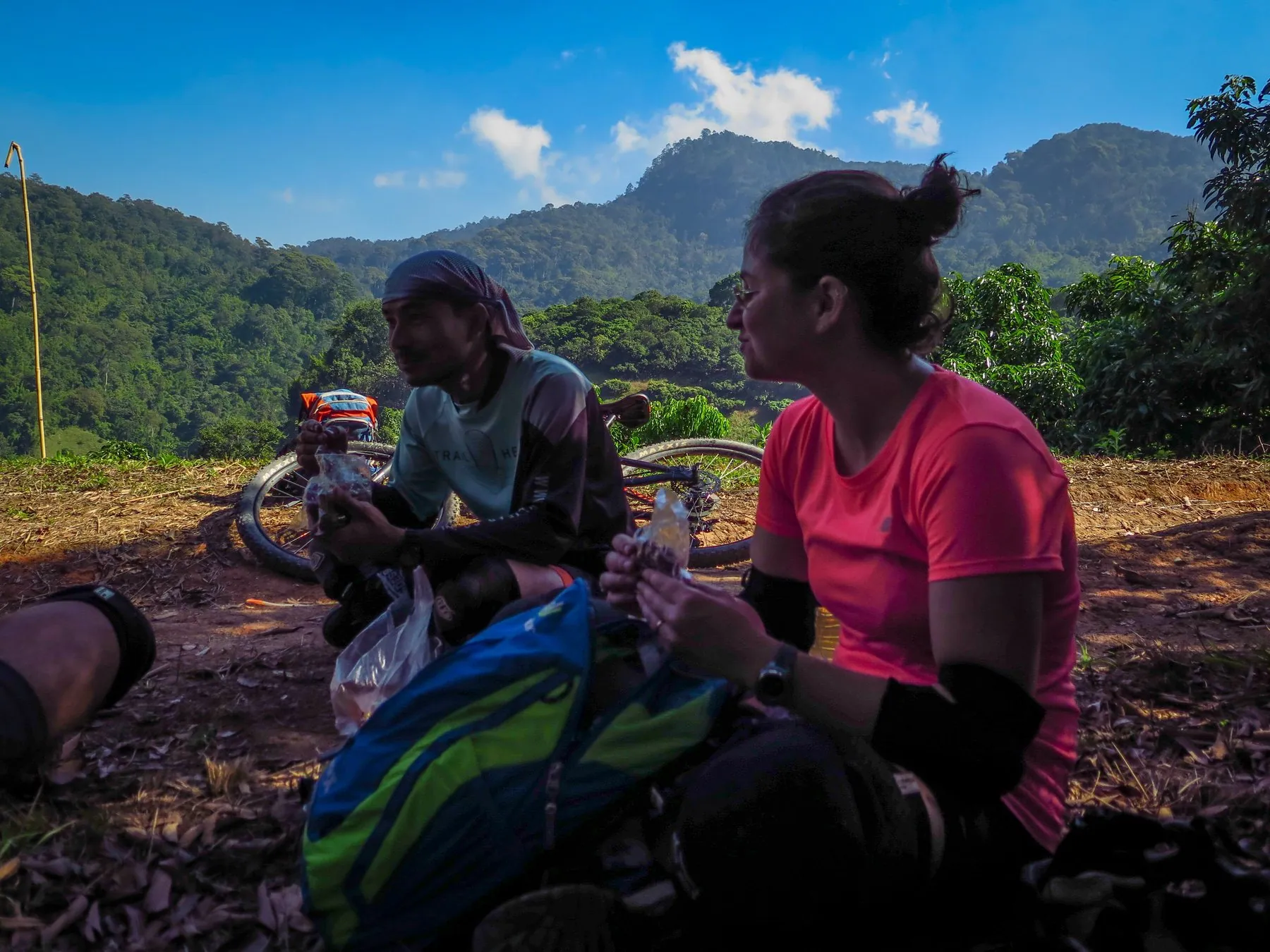 Two cyclists sitting on the ground beside their bikes taking a drink, a forested ridge rising behind them
