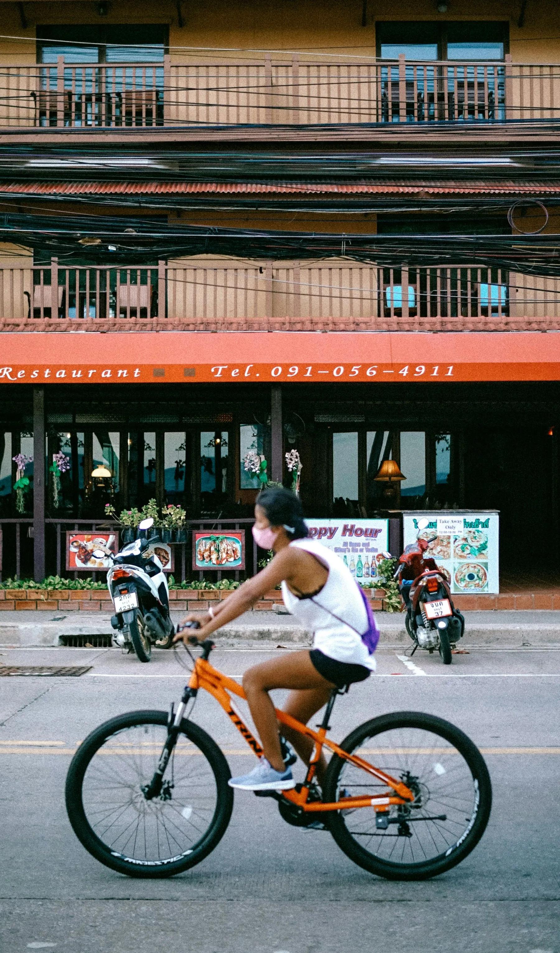 A man in a white shirt riding an orange mountain bike past a Thai shophouse with a red Thai-script sign and parked motorbikes