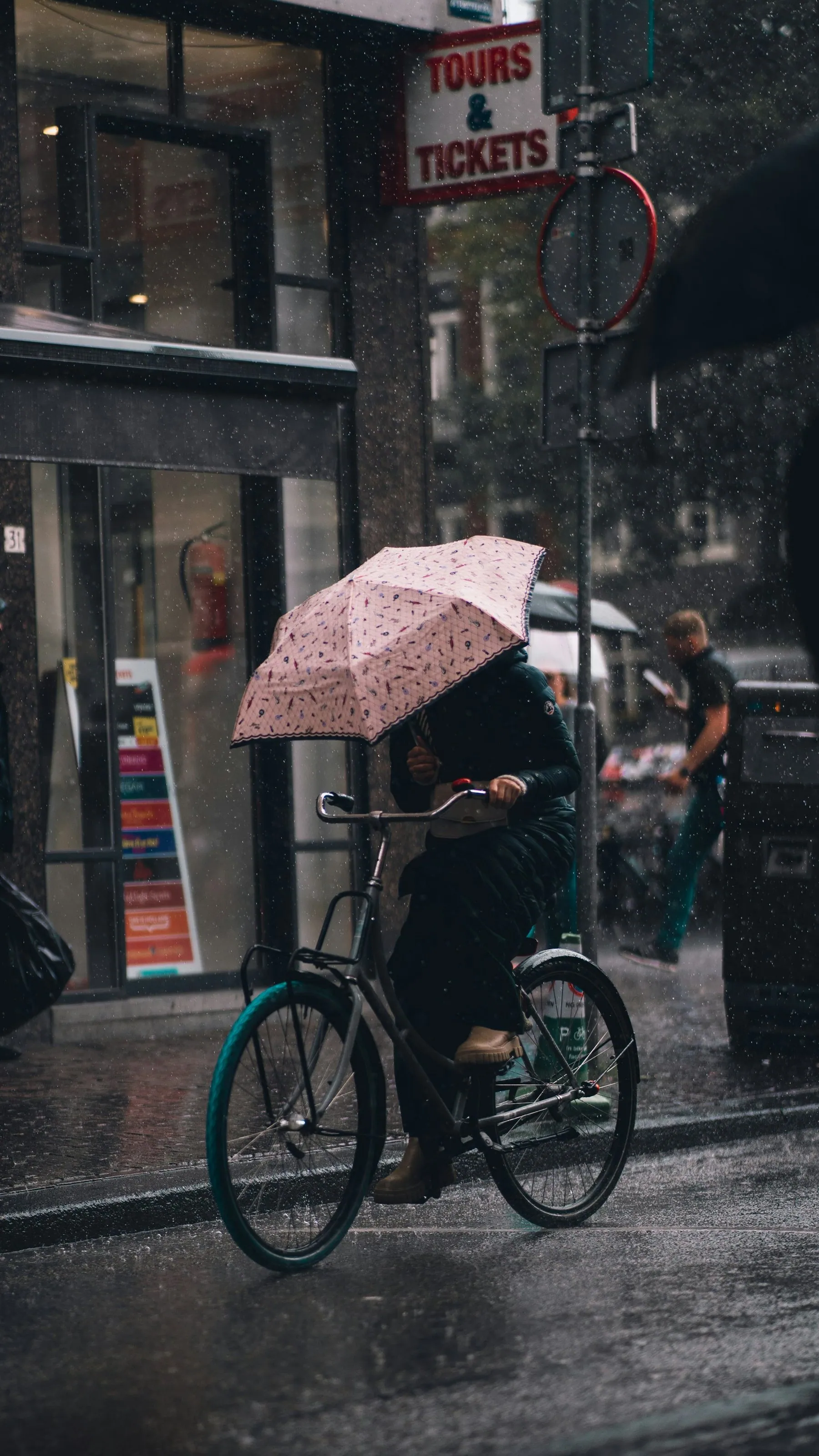 A cyclist riding one-handed under a pink umbrella through heavy rain past a tours and tickets shopfront