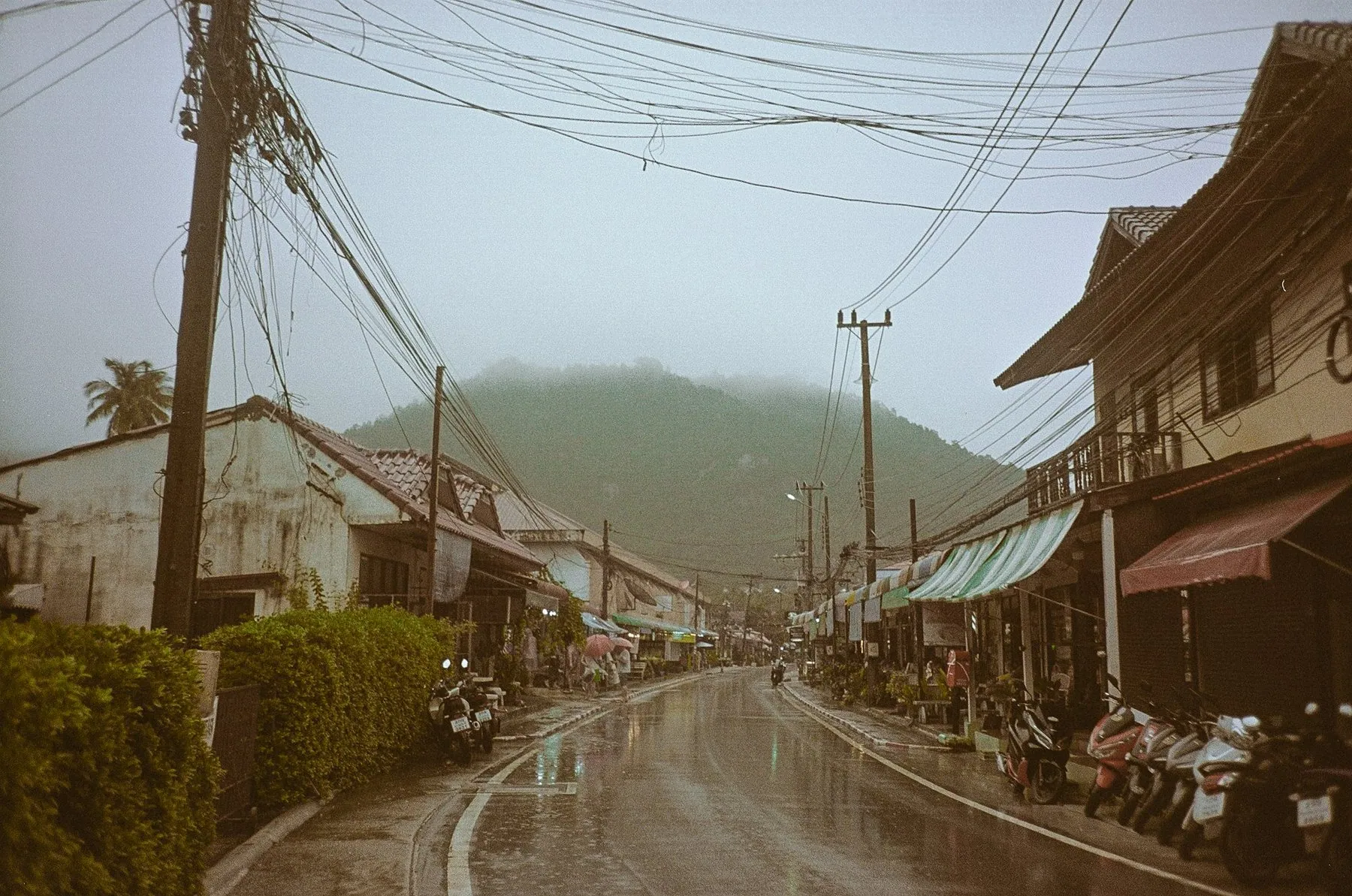 A rain-soaked street in a Thai hill town, scooters parked along both kerbs under tangled power lines with cloud sitting on the ridge behind