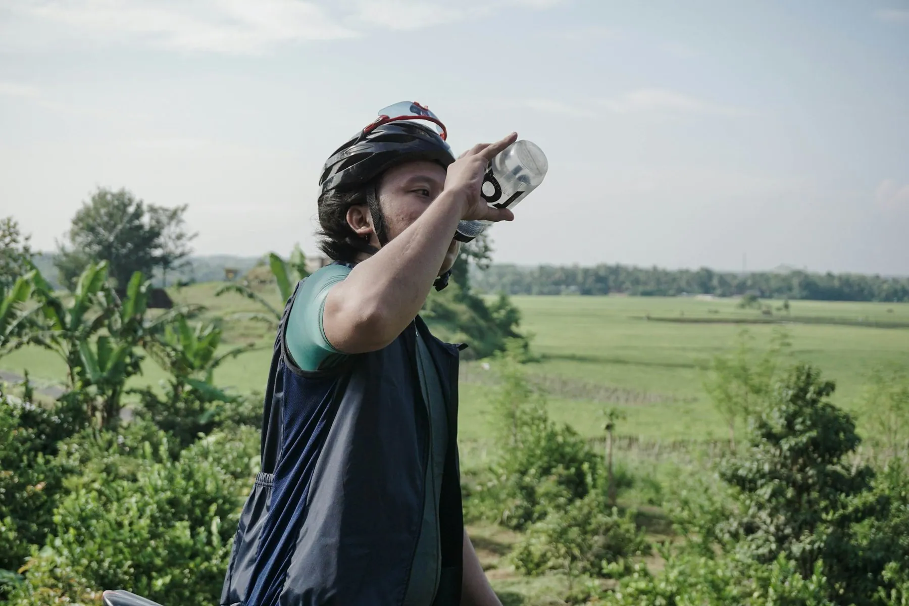 A man in a cycling helmet drinking from a bottle with flat green fields and palms behind him