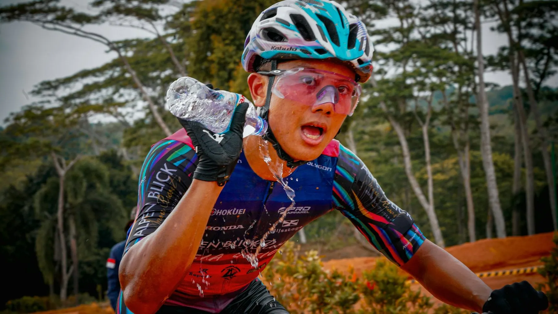 A cyclist in a helmet and sunglasses squeezing a water bottle over his face on a road through dense green forest
