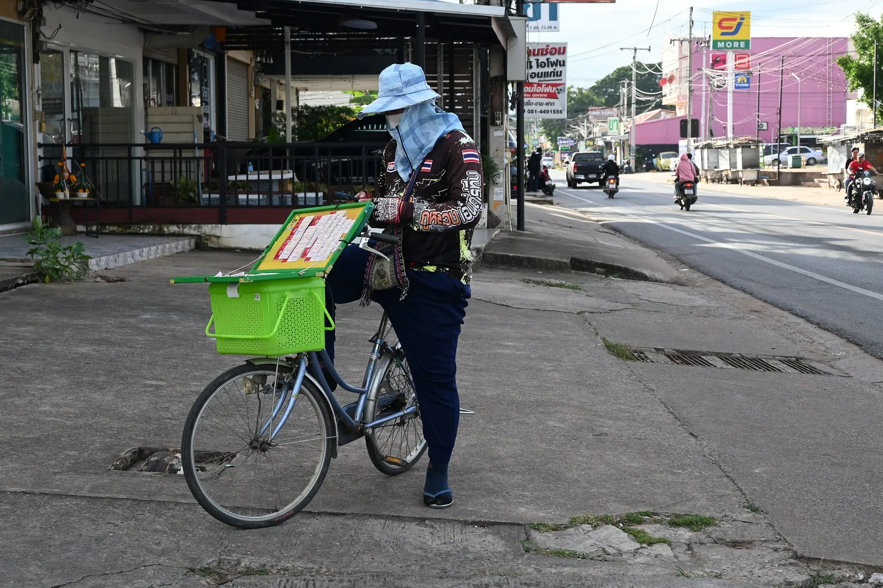 A vendor in a wide-brimmed hat stands with a loaded bicycle on a Thai pavement beside a road, a 7-Eleven sign behind