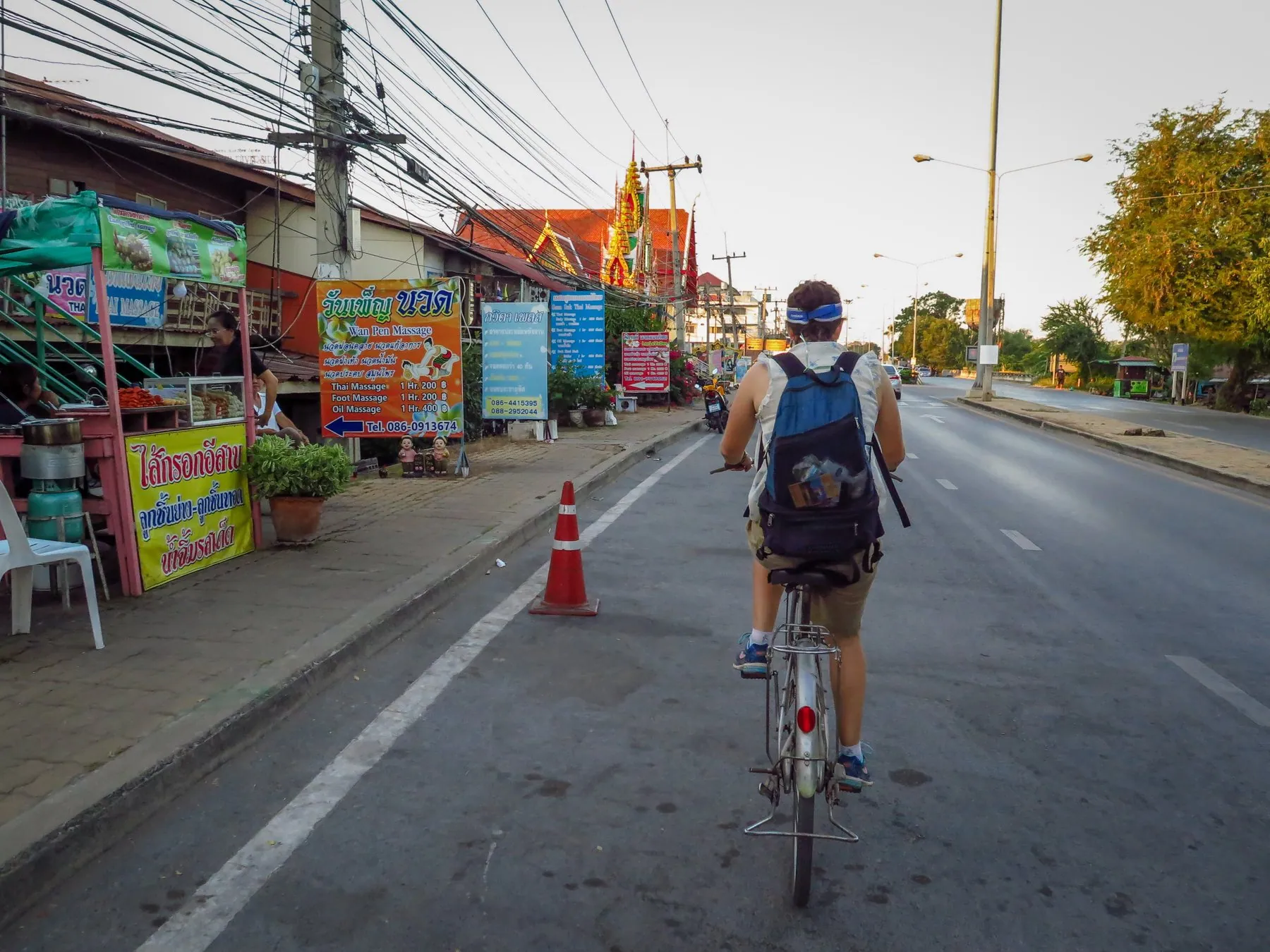 A cyclist seen from behind riding down the middle of a Thai street past shopfronts and a traffic cone