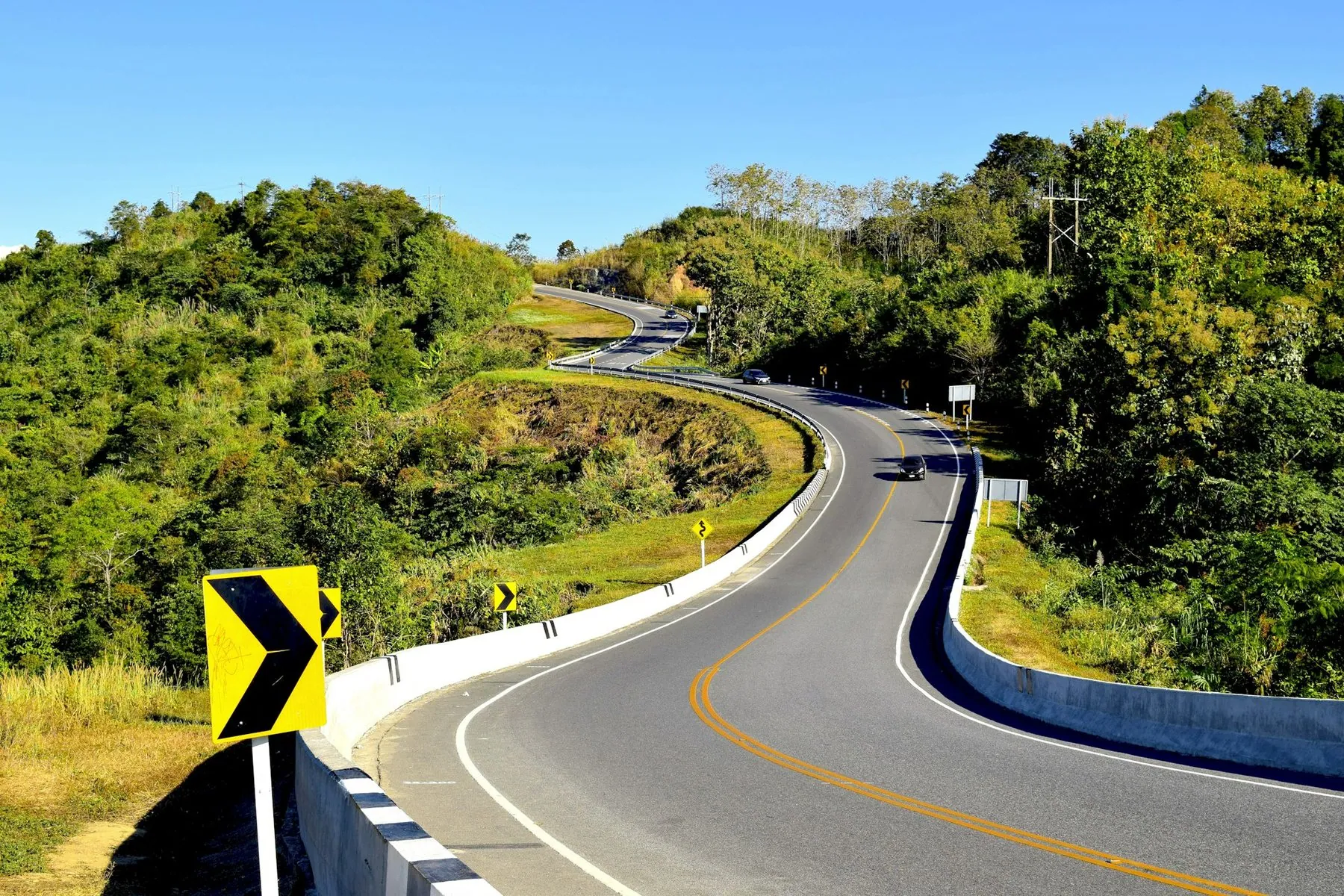 A tarmac road curving left around a wooded hillside with a yellow chevron sign on the outside of the bend