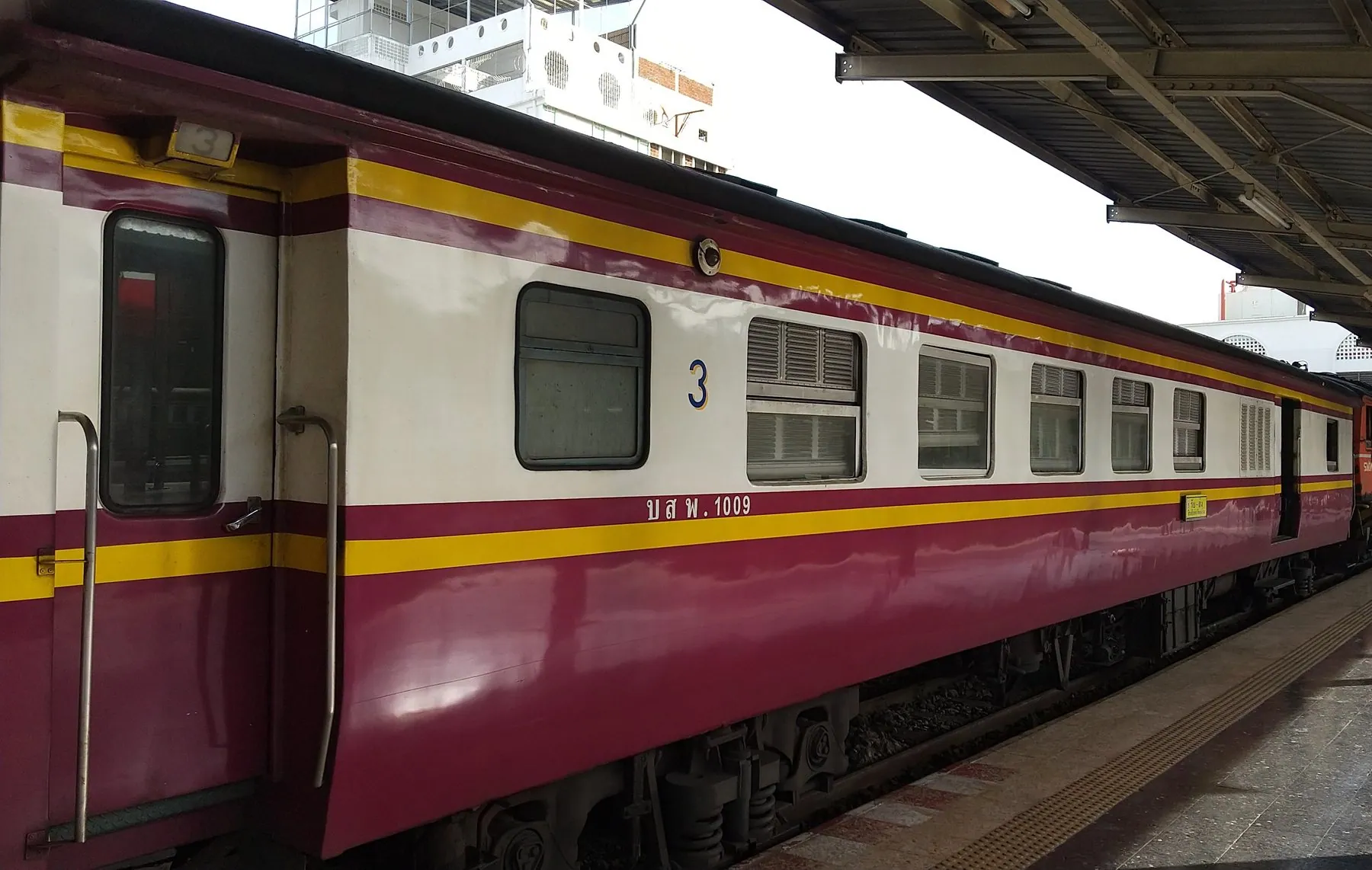 A maroon and cream third-class State Railway of Thailand carriage standing at a platform in Bangkok with its end door open