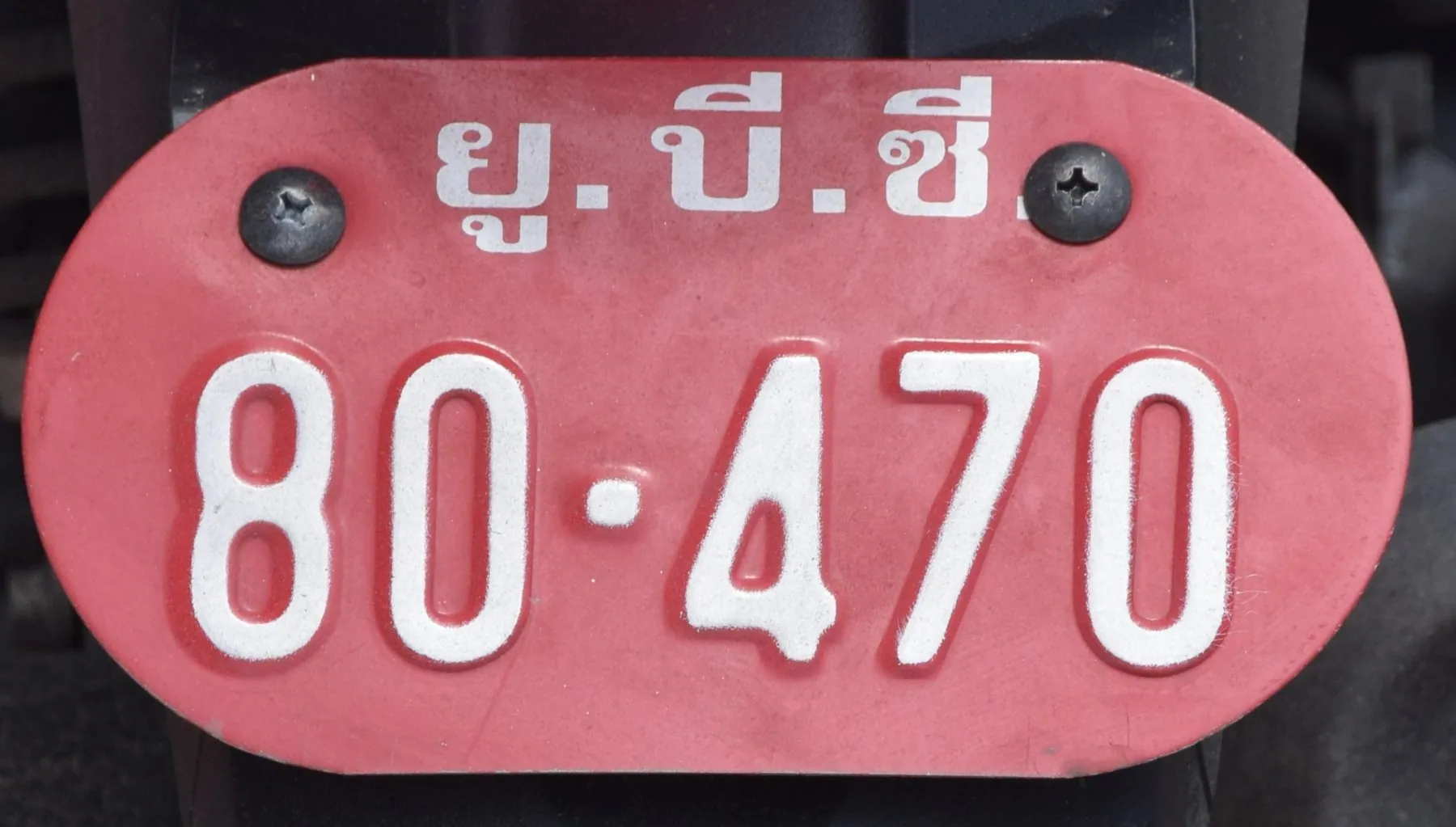 A small oval red motorcycle plate bolted to a Thai moped, the digits in white