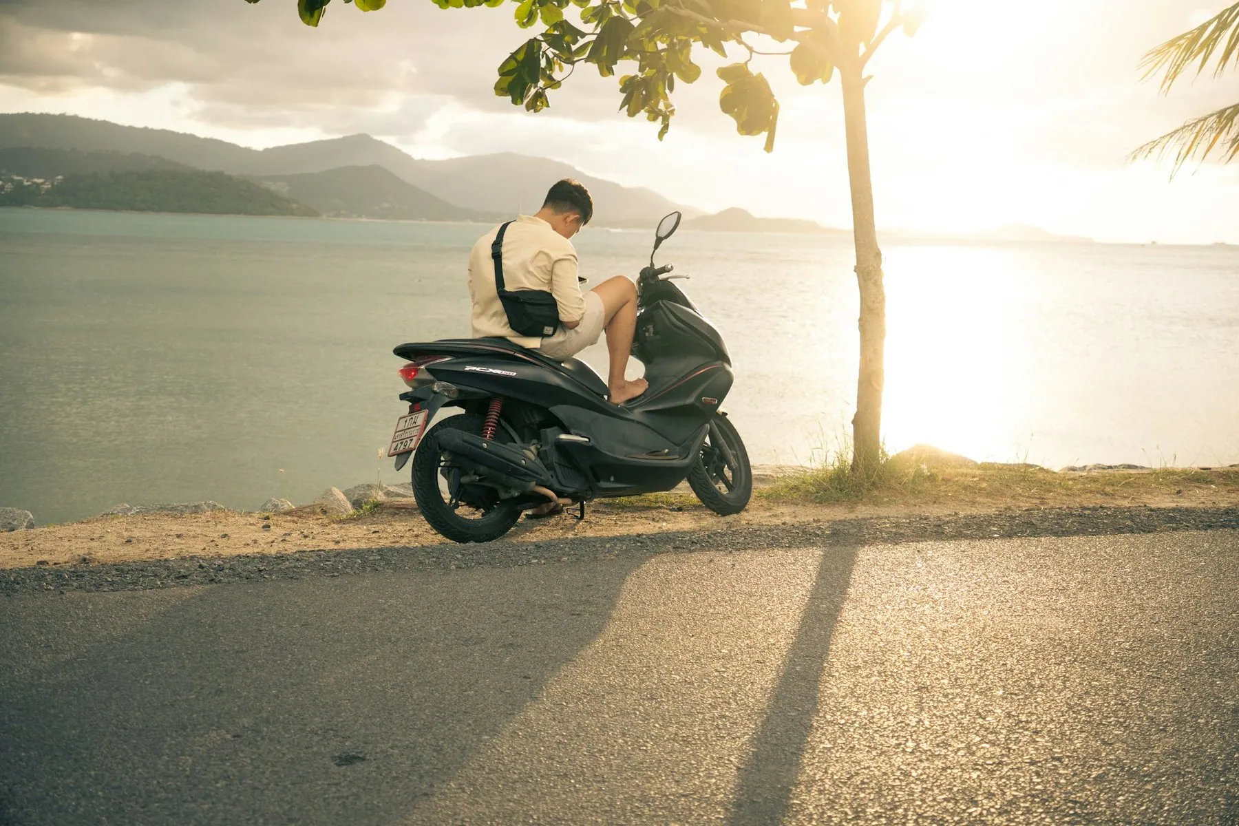 A man sitting on a parked scooter at the roadside looking out over the sea at sunset