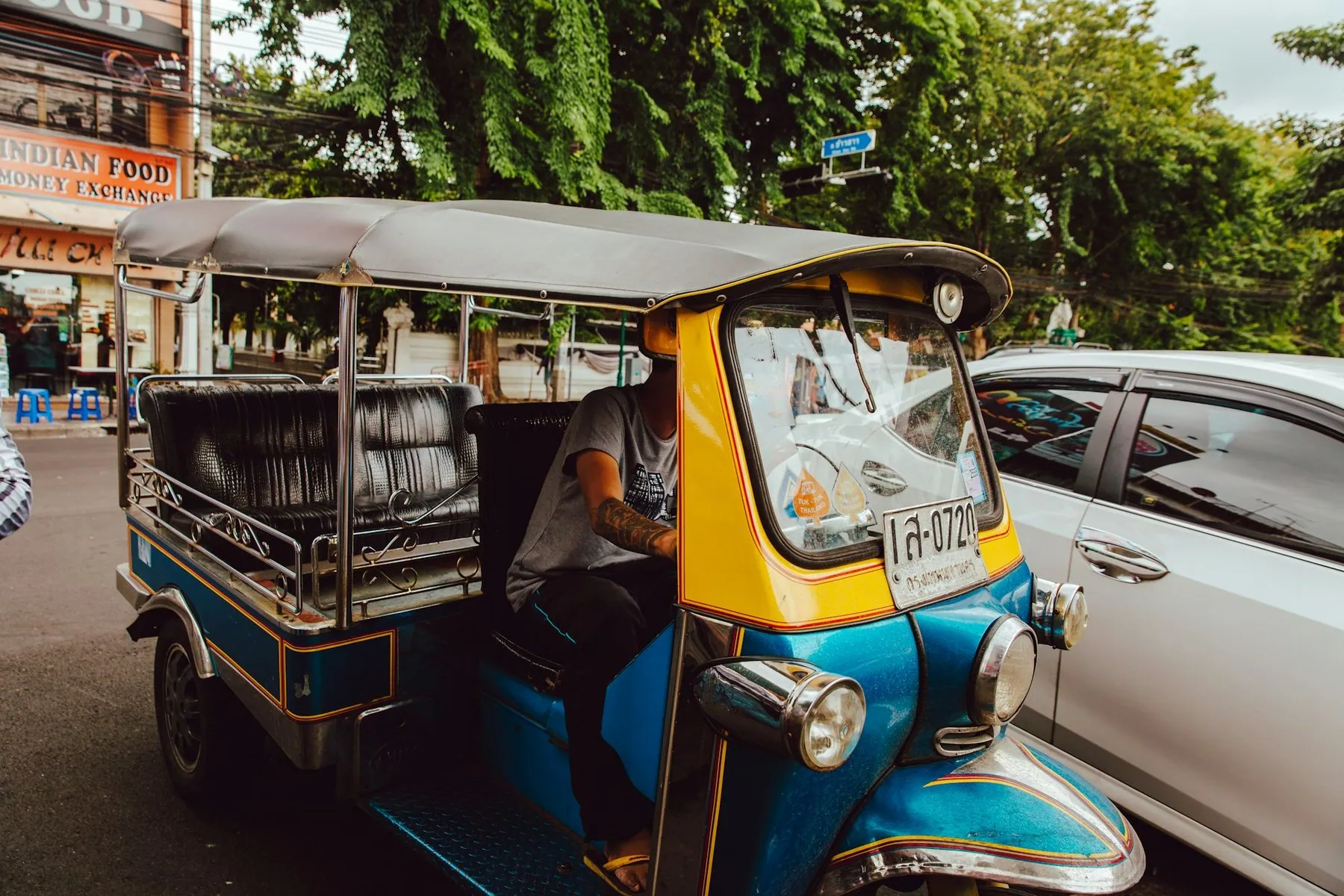 A blue and yellow tuk-tuk with a Thai number plate waiting in traffic beside a white car
