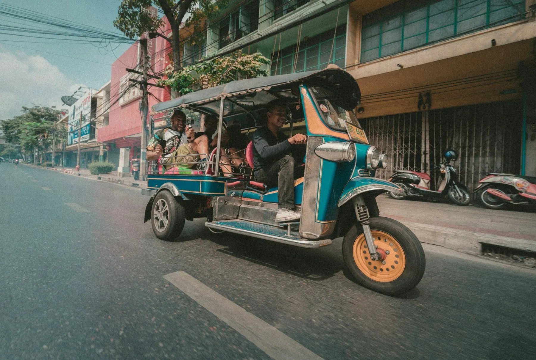 A green and blue tuk-tuk carrying passengers along a wide, quiet Thai street