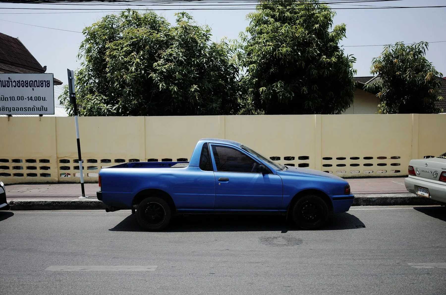 A plain blue pickup parked at the kerb in front of a wall carrying a Thai-script noticeboard