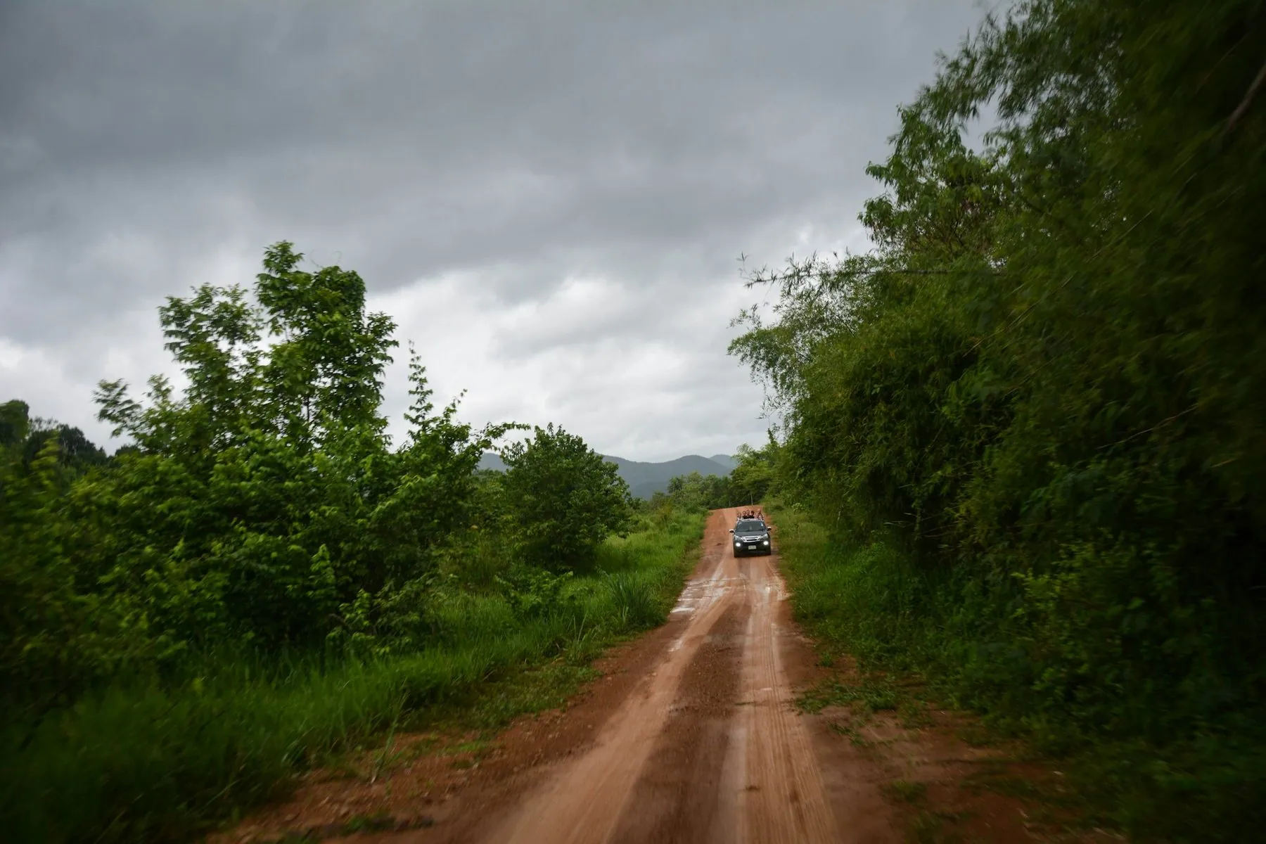 A car far down a red dirt track running between walls of tropical greenery under grey cloud