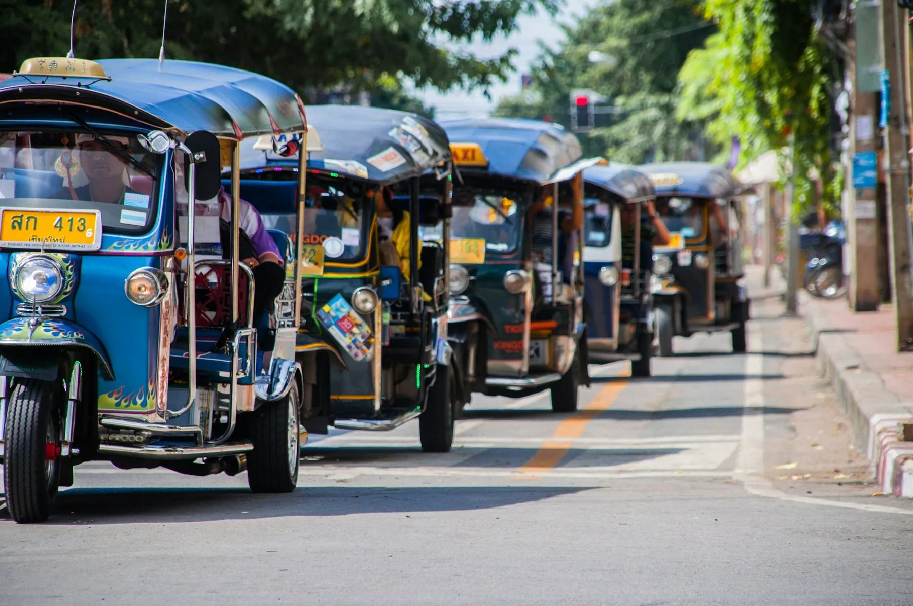A line of blue and yellow Thai tuk-tuks waiting nose to tail at the roadside, number plates showing