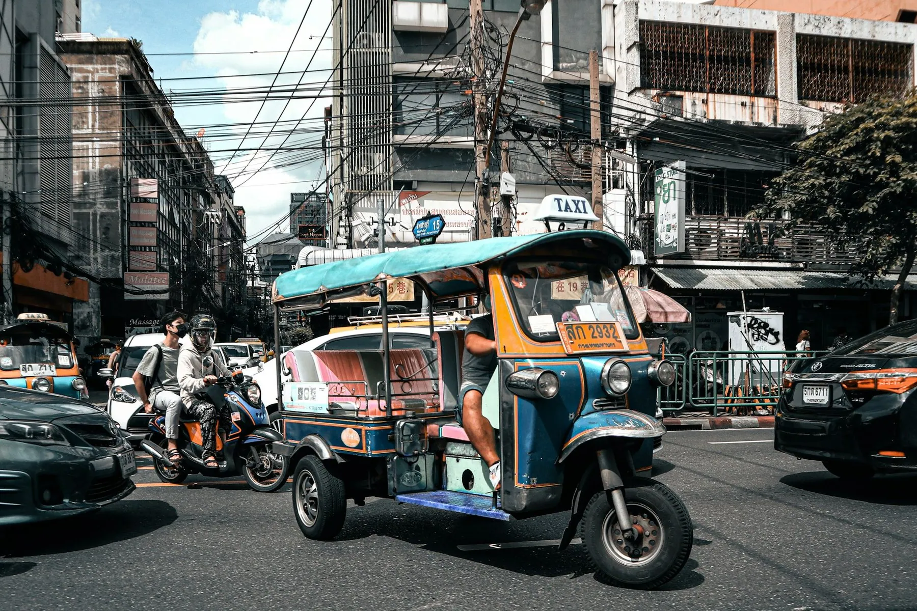 Scooters and a blue tuk-tuk waiting side by side at a busy junction under Bangkok power lines