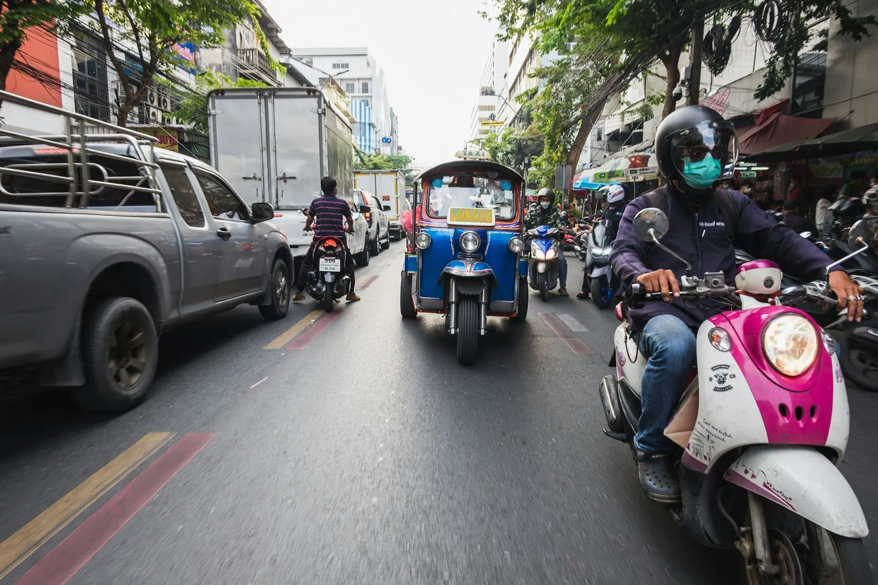 A tuk-tuk, a pickup and several scooters moving together down a Bangkok street, seen from behind
