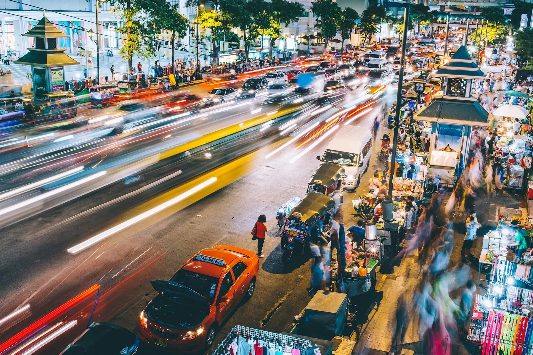 Long-exposure light trails from cars and motorbikes crossing a wide Bangkok intersection at dusk