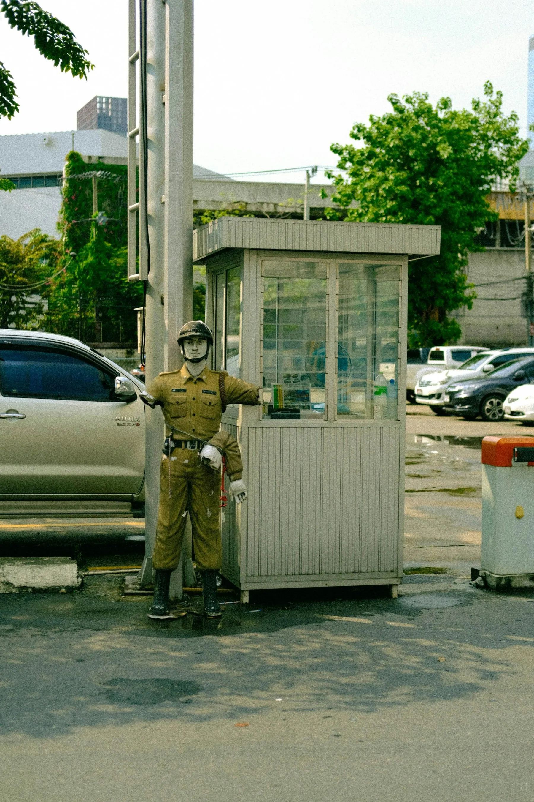 A uniformed attendant standing beside a small white sentry booth at the entrance to a Thai compound