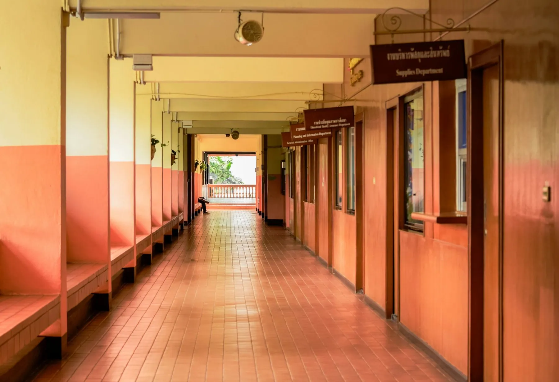 A long corridor of a Thai public building lined with wooden benches and Thai-script department signs