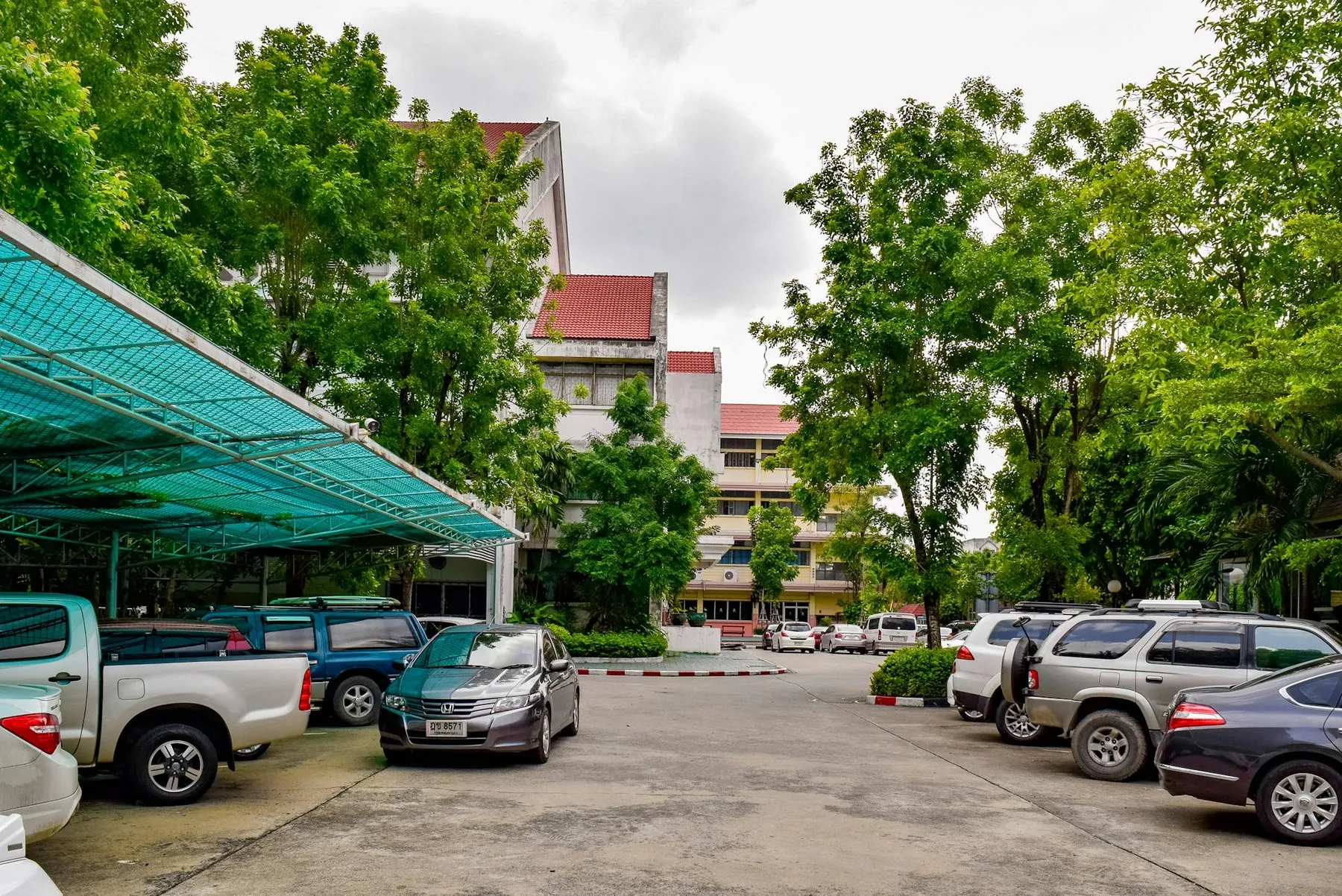 Pickups and cars parked under a green canopy outside a Thai public building