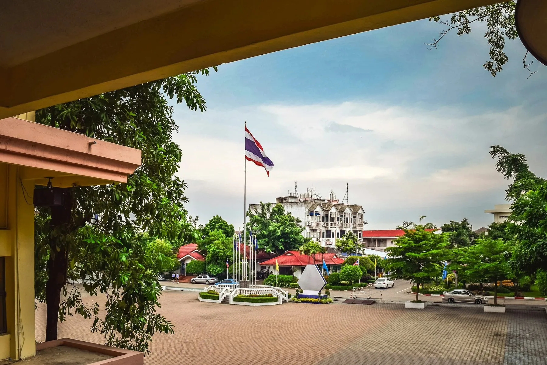 A Thai flag flying over a low government building seen from a shaded walkway