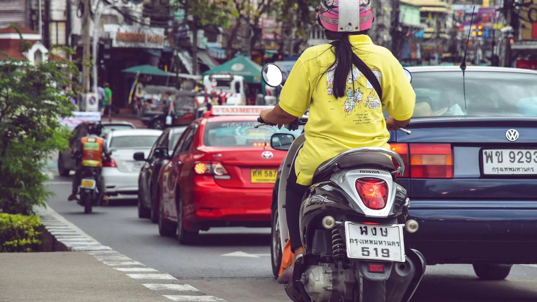 A rider in a yellow shirt and pink helmet threading a scooter between cars in Bangkok traffic
