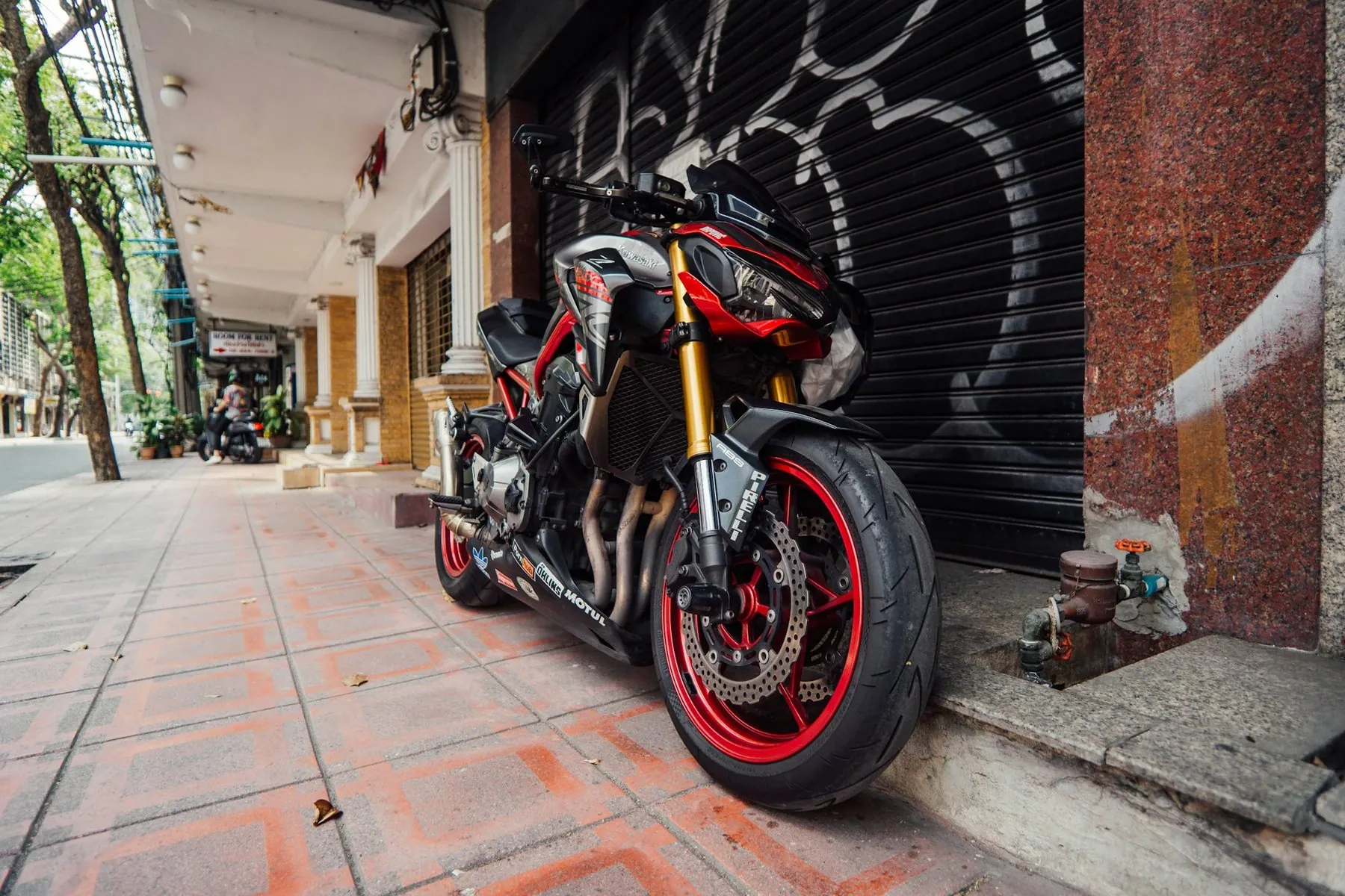 A large red and black naked motorcycle parked on a tiled pavement in front of a shuttered shop