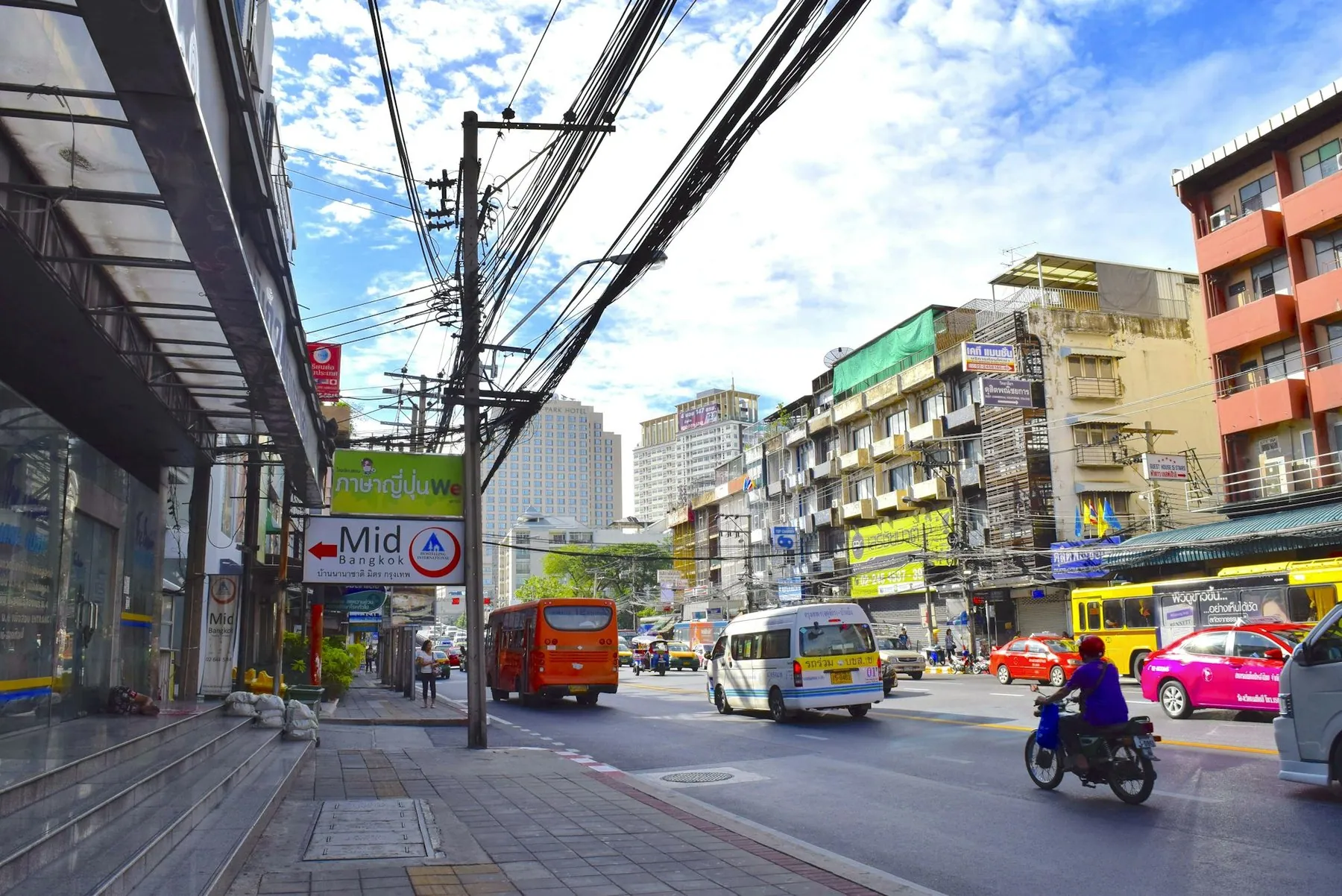 A Thai street seen from the pavement, with a minibus, a scooter and tangled overhead cables