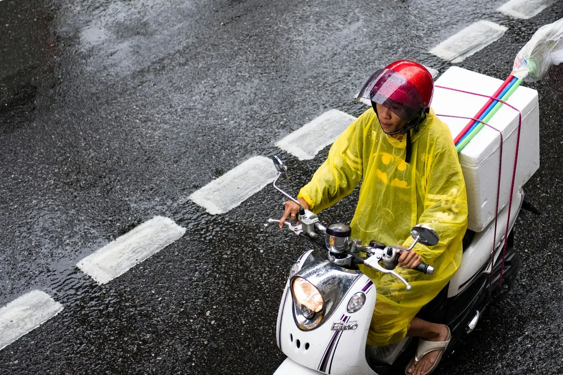 Seen from directly above, a rider in a yellow rain jacket on a scooter loaded with a white delivery box