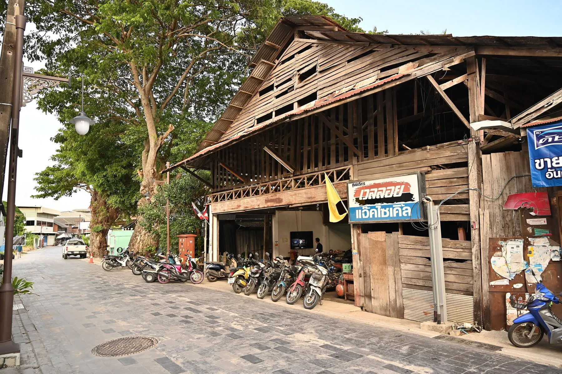 Motorbikes lined up along the front of a weathered wooden shophouse on a quiet sunlit street, a Thai motorcycle-shop sign hanging above them