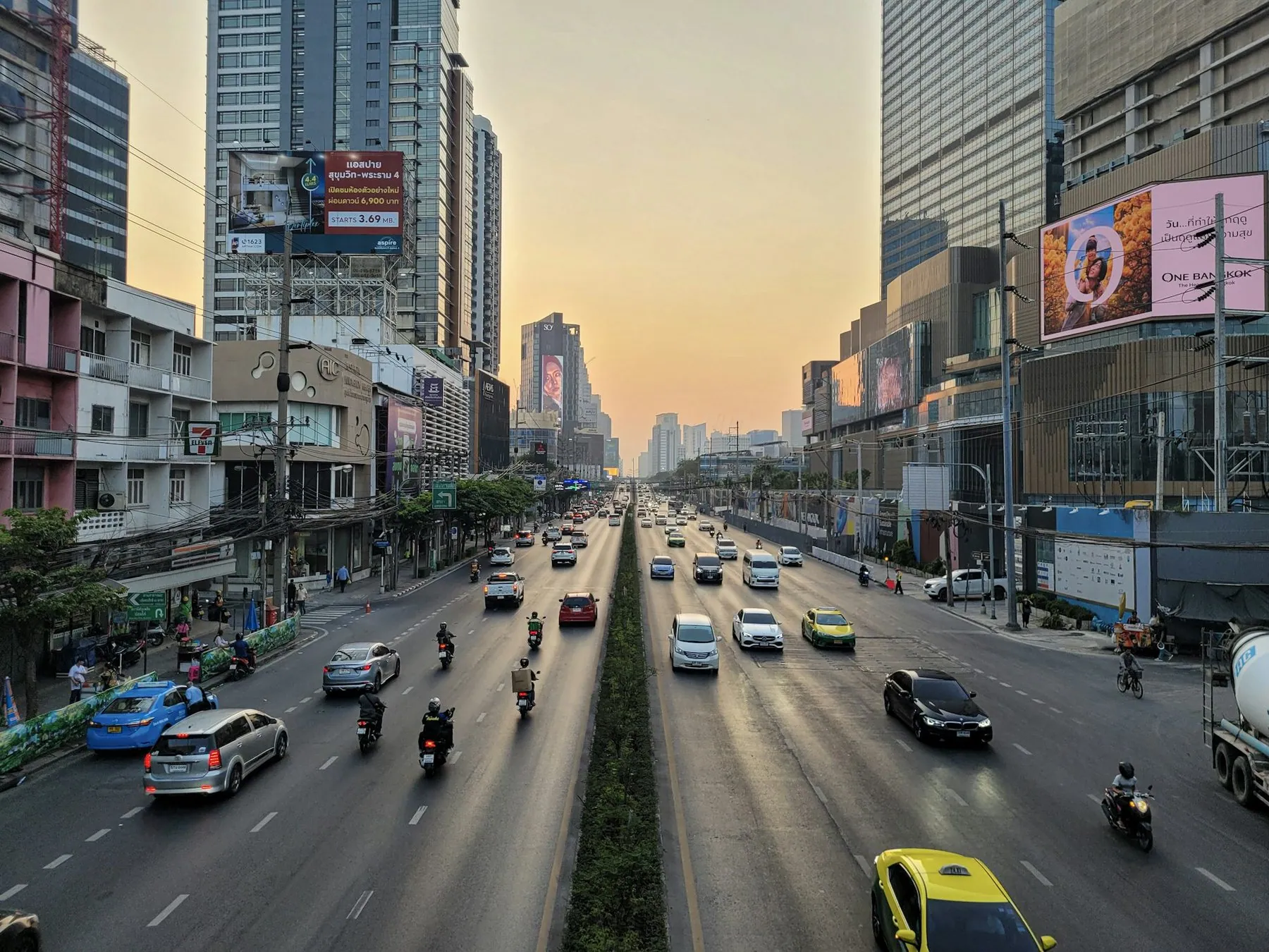 A wide Bangkok avenue at dusk with cars, motorbikes and a yellow-green taxi between tower blocks