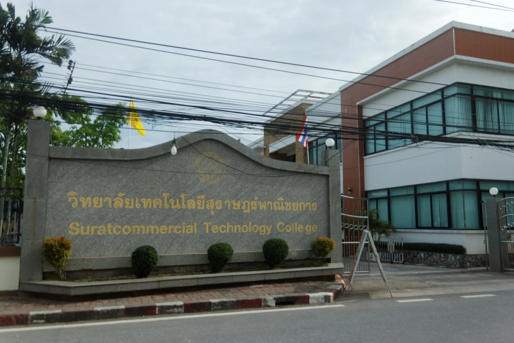 A wide street through Surat Thani town past a college wall, power lines overhead