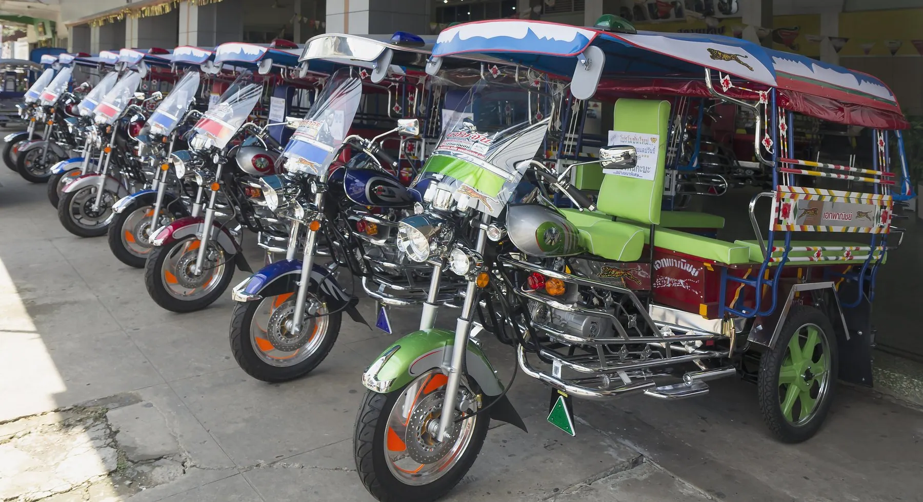 A line of new Isaan-style three-wheeler tuk-tuks and motorbikes for sale on a forecourt in Udon Thani