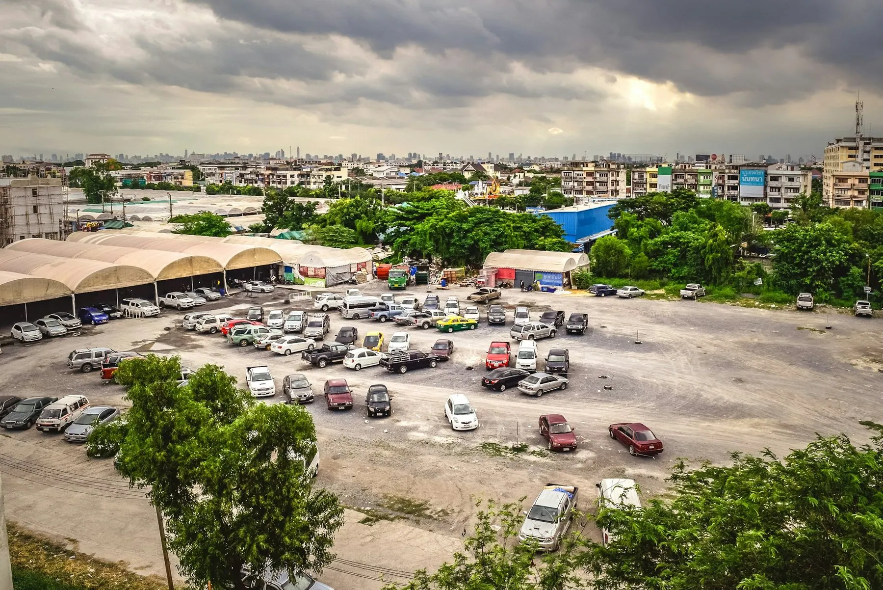 An open gravel car park in Bangkok filled with pickups and saloons, a green and yellow taxi among them and long metal canopies along one side