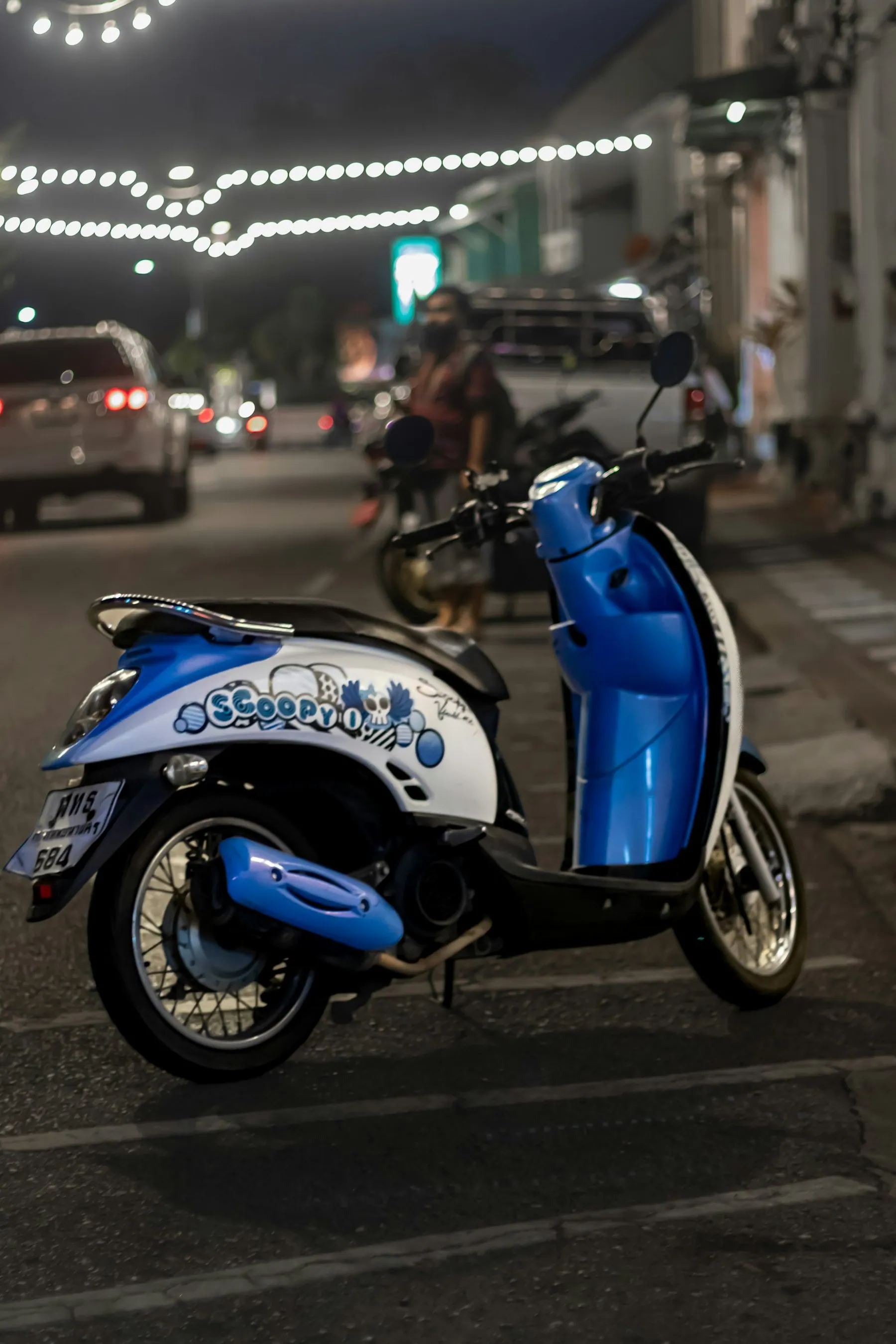 A blue and white retro-styled automatic scooter parked at the kerb under evening lights