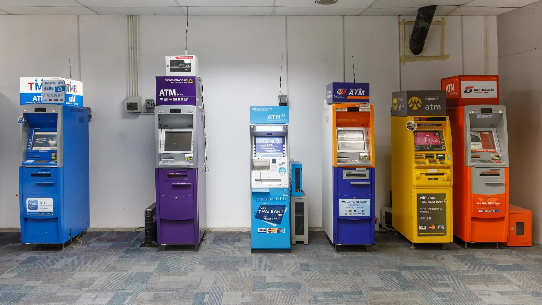 A line of blue, purple, green, yellow and orange Thai bank cash machines standing against a wall inside Phuket International Airport