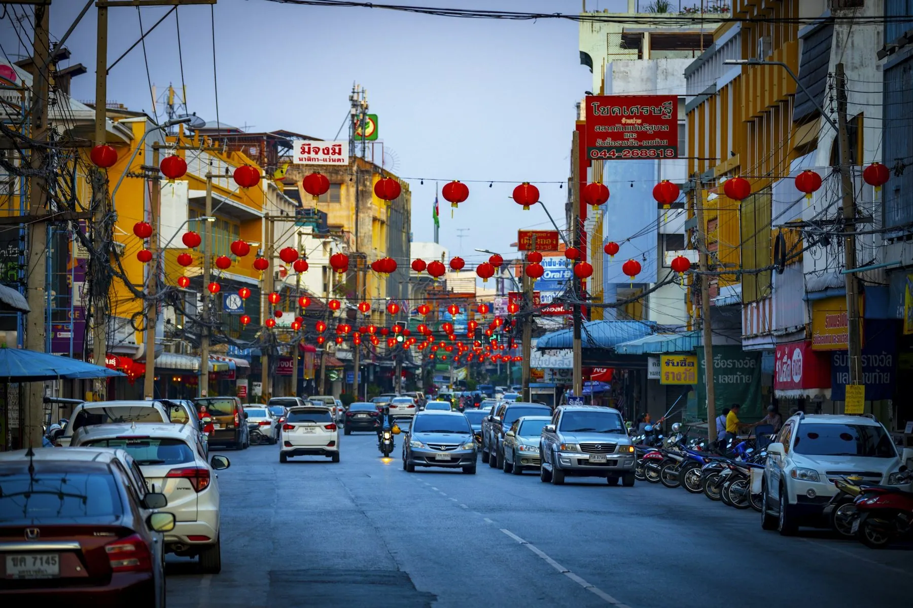 Cars and motorbikes parked along both sides of a Thai shophouse street strung with red lanterns